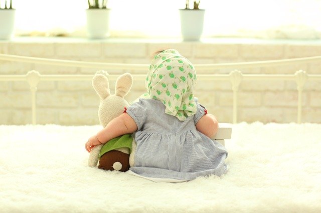 Bábätko v šatách a šatke na hlave sedí na posteli s plyšovým zajacom.jpg